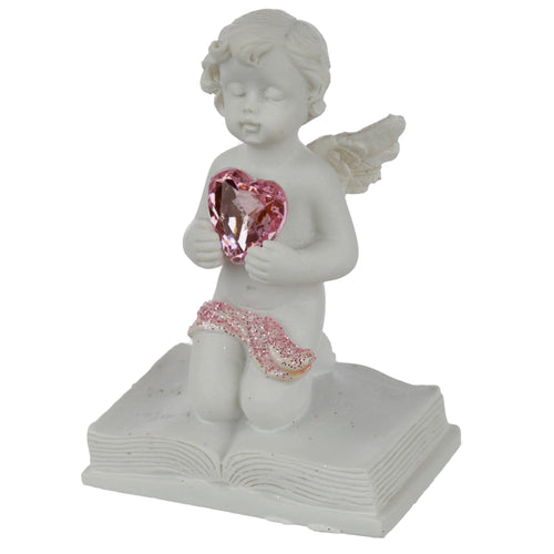 Angled view of a white cherub figurine kneeling on an open book. It holds a faceted pink heart jewel to its chest and features closed eyes, small wings, and a pink glitter sash.