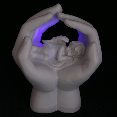 White cherub figurine with wings sleeping in cupped hands, illuminated by a purple LED light; angled view against a dark background.