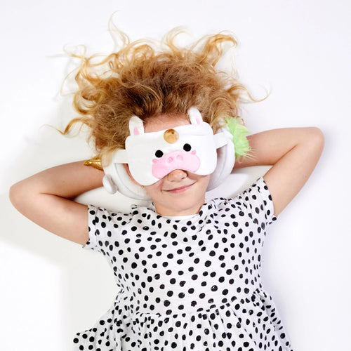In-context lifestyle shot showing a child lying down, wearing a white unicorn eye mask with a golden horn, pink snout, and ears, alongside a white plush travel pillow with green fluffy detail.