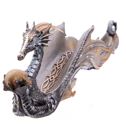 Angled view of a shimmering metallic grey and gold dragon-shaped incense ashcatcher. The dragon, with red eyes, is depicted holding a light brown skull in its claw. Its detailed body forms the sides of the patterned ash-catching trough, with an incense stick holder at the far end.