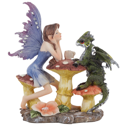 Woodland Spirit Fairy and Dragon Tea Party ornament. A brown-haired fairy in a blue dress with shimmering purple wings sits on a red toadstool, looking at a green dragon with red eyes on another toadstool. A teacup is on the mushroom table between them, on a textured brown base with a pink flower.