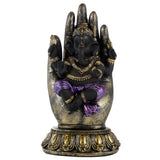 Front view of a black Ganesha figurine, adorned with gold jewellery and a purple dhoti, seated within a textured gold and black hand ornament, resting on an ornate gold base.