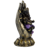 Angled view of a purple, gold, and black Ganesh in Hand figurine ornament. The black Ganesh, with gold details and purple attire, sits within a large, textured gold and black hand sculpture, resting on an ornate matching base.