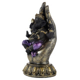 Angled view of a black Ganesh figurine with purple and gold detailing, seated within a large gold and black hand, set on an ornate gold and black base.