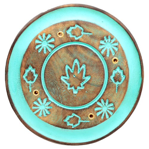 Overhead view of a round wooden incense holder, depicting a central turquoise painted leaf design surrounded by smaller leaf and starburst patterns. The ashcatcher features brass-coloured holes for incense sticks and a turquoise outer border.