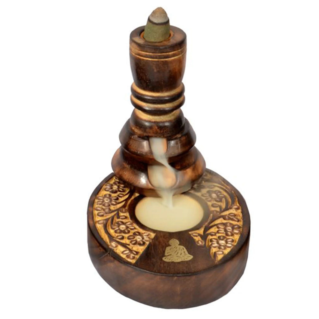 Angled view of a dark carved mango wood backflow incense burner, showing white smoke flowing from the top cone into the round base with intricate golden floral patterns and a meditating Buddha figure.