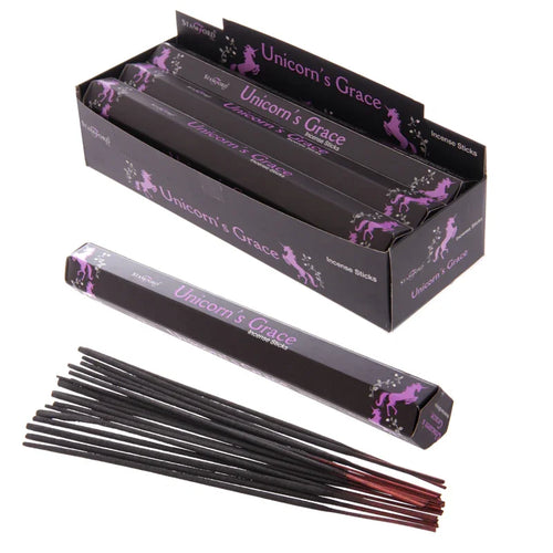 Angled view of a black display box filled with multiple black incense stick packs, each bearing 'Unicorn's Grace' text and purple unicorn illustrations. In the foreground, a single black 'Unicorn's Grace' incense stick pack is placed beside a cluster of dark, slender incense sticks with reddish ends.