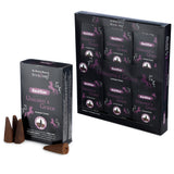 Angled view of black and purple 'Unicorn's Grace' backflow incense cone packaging featuring unicorn graphics. A multipack display box, containing six smaller packs, is positioned behind an individual pack. Several brown conical backflow incense cones, one showing its hollow base, are scattered in front.