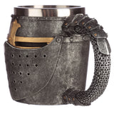 Front-angled view of a decorative medieval helmet tankard, showing its textured dark grey, distressed finish. The helmet design features a golden cross on the visor, horizontal eye slits, and ventilation holes. The tankard handle resembles chain mail and armour plates.