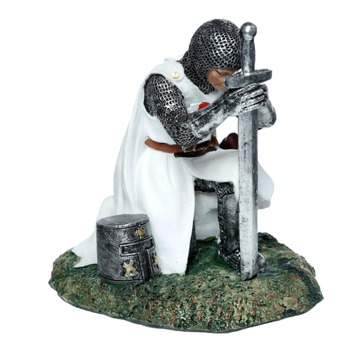 Kneeling knight figurine in silver-coloured chainmail and white surcoat with a red cross, holding an upright sword, with a great helm beside him on a textured green base, shown from an angled perspective.