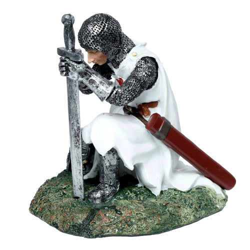 Angled view of a kneeling knight ornament, wearing silver chainmail armour and a white surcoat with a red cross, holding an upright sword, on a textured green base.