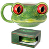 A ceramic tree frog head mug, primarily green with mottled markings and striking red and black eyes, is shown from an angled perspective with its handle visible. Beneath it, the dark green product box, labelled 'TREE FROG SHAPED HEAD MUG' and patterned with red and green zigzags, displays a portion of the mug through a cut-out.