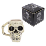 Front view of an off-white, distressed ancient skull head ceramic shaped mug with black eye sockets and a bone-shaped handle, displayed alongside its black box.