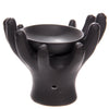 Angled view of a black ceramic oil burner, designed as open hands cupping a bowl, with a tealight opening visible at the base.