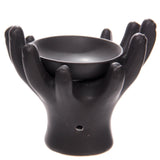 Angled view of a black ceramic oil burner, designed as open hands cupping a bowl, with a tealight opening visible at the base.