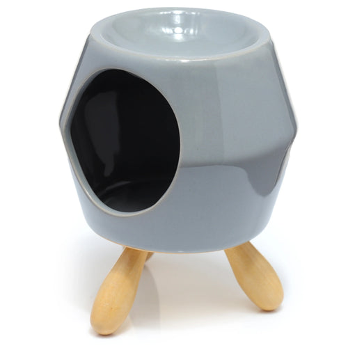 Angled view of an abstract, faceted grey ceramic oil burner, showing its recessed top bowl, circular side opening, and three natural wooden feet.