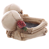 Angled view of a cream-coloured skull ashtray depicting two embracing skulls and a red rose, showing the dark textured interior.