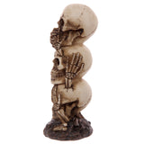 Angled view of a three-tiered bone-coloured skull totem decoration, each skull depicting 'See No, Speak No, Hear No Evil' with skeletal hands covering its eyes, mouth, or ears. The figurine stands on a dark, textured base.
