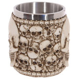 Angled view of a cream-coloured decorative tankard. Its exterior features a relief of multiple skulls and a bone-like border. It has a metallic silver interior lining, and no handle is visible.