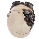 Angled view of a cream-coloured steampunk skull ornament featuring dark goggles on its crown and intricate metallic gears visible on its fractured side, against a plain white background.