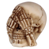 Angled close-up of an aged cream and brown 'See No Evil' skull figurine, with skeletal hands covering its eye sockets.