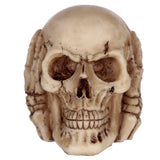 Front view of an aged, bone-coloured skull ornament with hands covering its eyes, in a 'See No Evil' pose.