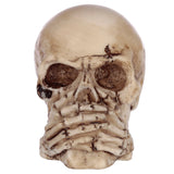 Close-up, front view of an aged, bone-coloured skull ornament, depicting the 'Speak No Evil' pose with hands covering its mouth and dark, textured details.