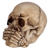 Angled close-up of a small, bone-coloured skull ornament with skeletal hands covering its mouth, depicting the 'Speak No Evil' pose.