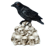 Decorative black crow ornament with textured plumage and red eyes, standing on a pile of bone-coloured skulls, depicted from an angle.