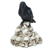 Front view of a black crow ornament with red eyes and textured feathers, perched on a pile of aged, cream-coloured skulls.