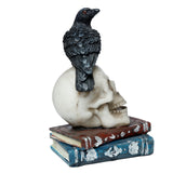 Angled view of a black crow figurine with orange eyes, perched on an off-white skull resting on two stacked ornate books, one red-brown and one blue, on a white background.