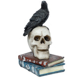Front view of a black crow figurine perched on an off-white skull, resting on a stack of red and blue antique-style books with silver detailing, on a white background.