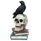 Angled view of a black crow ornament with red eyes perched atop a cream-coloured skull, which sits on two stacked, distressed books in red-brown and blue hues.
