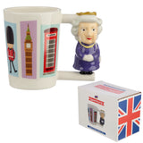 Angled view of a white ceramic mug featuring a Queen figure handle in a purple dress and gold crown. The mug's body is decorated with colourful illustrations of a Beefeater, Big Ben, and a red telephone box. A white gift box with a Union Jack design is visible.
