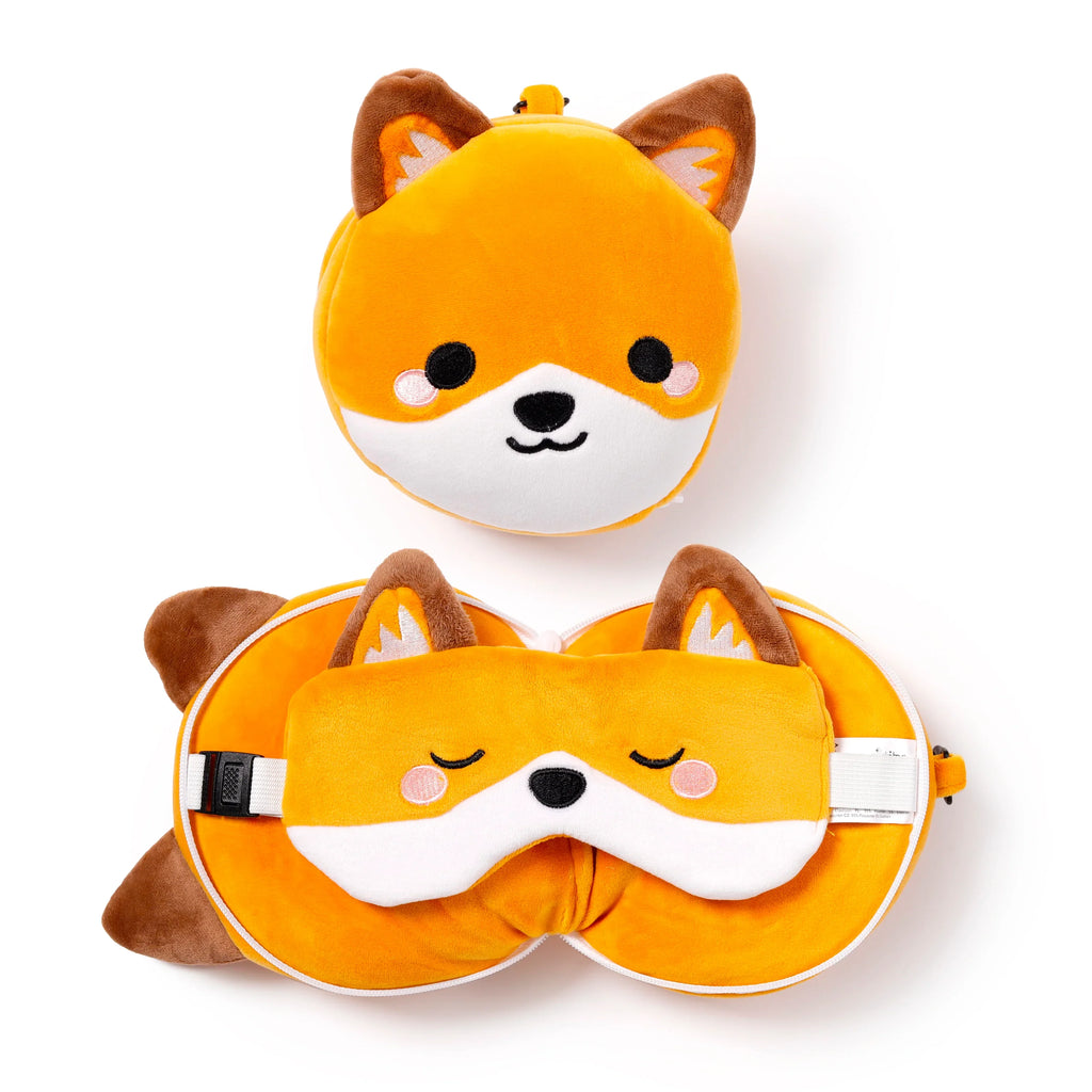 Overhead view of an orange and brown fox travel pillow and eye mask set on a white background. The set includes a compact, folded fox head pillow with black eyes and nose, and an unfolded U-shaped travel pillow with a matching fox eye mask featuring closed eyes and a black nose, secured by a white strap and buckle.