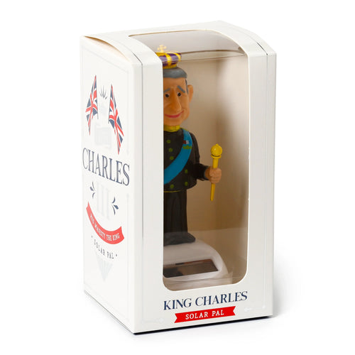 Angled front view of a collectible King Charles solar powered pal figure, depicted wearing a crown and royal uniform, visible through the clear plastic window of its white display box. The packaging displays 'King Charles Solar Pal' text and Union Jack flags.