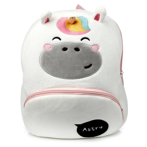 Front view of a white plush unicorn backpack featuring a gold horn, rainbow mane, and embroidered unicorn face with pink cheeks. It has pink zippers and the name Astra embroidered on a black patch.
