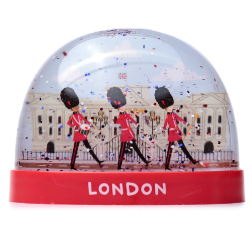 Front view of a collectable London snow globe featuring three marching Queen's Guards in front of Buckingham Palace with a Union Jack flag, and red, white, and blue glitter. The red base displays 'LONDON' in white text.