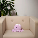 Front view of a lilac Plush Squeezies octopus cushion with white spots and a smiling face, sitting on a beige armchair.