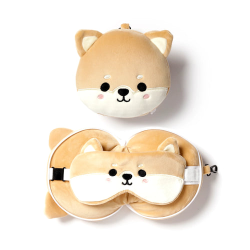 Flat lay showing a tan and cream plush Shiba Inu travel pillow and eye mask set on a white background. At the top is the compact, round Shiba Inu head. Below it, the U-shaped pillow is open, with the matching Shiba Inu eye mask resting on its surface, showing an adjustable strap with a black buckle.
