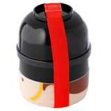 Angled view of a round, stacked bento lunch box. The design features a black domed top resembling a bearskin hat, with a beige lower section depicting a London Guardsman's face. A bright red elastic strap secures the stacked compartments.