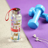 In-context lifestyle shot of a clear water bottle, standing upright on a grey surface, featuring a colourful wrap-around design of London icons and a metallic lid with a red carrying loop. In the background, a purple exercise mat displays blurred blue dumbbells and white earphones.