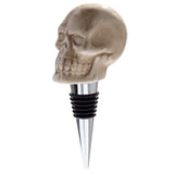 Angled view of a beige ceramic skull bottle stopper with a glossy finish, showcasing detailed facial features and a silver-coloured metal base with black rubber rings, on a white background.