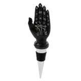 Front view of a black tarot hand bottle stopper featuring white astrological symbols on the palm and fingers, with a silver and black rubber stopper.