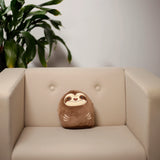 Lifestyle shot of a brown Plush Squeezies Sloth Cushion, facing forward with a smiling face and embroidered claws, sitting on a soft cream armchair.