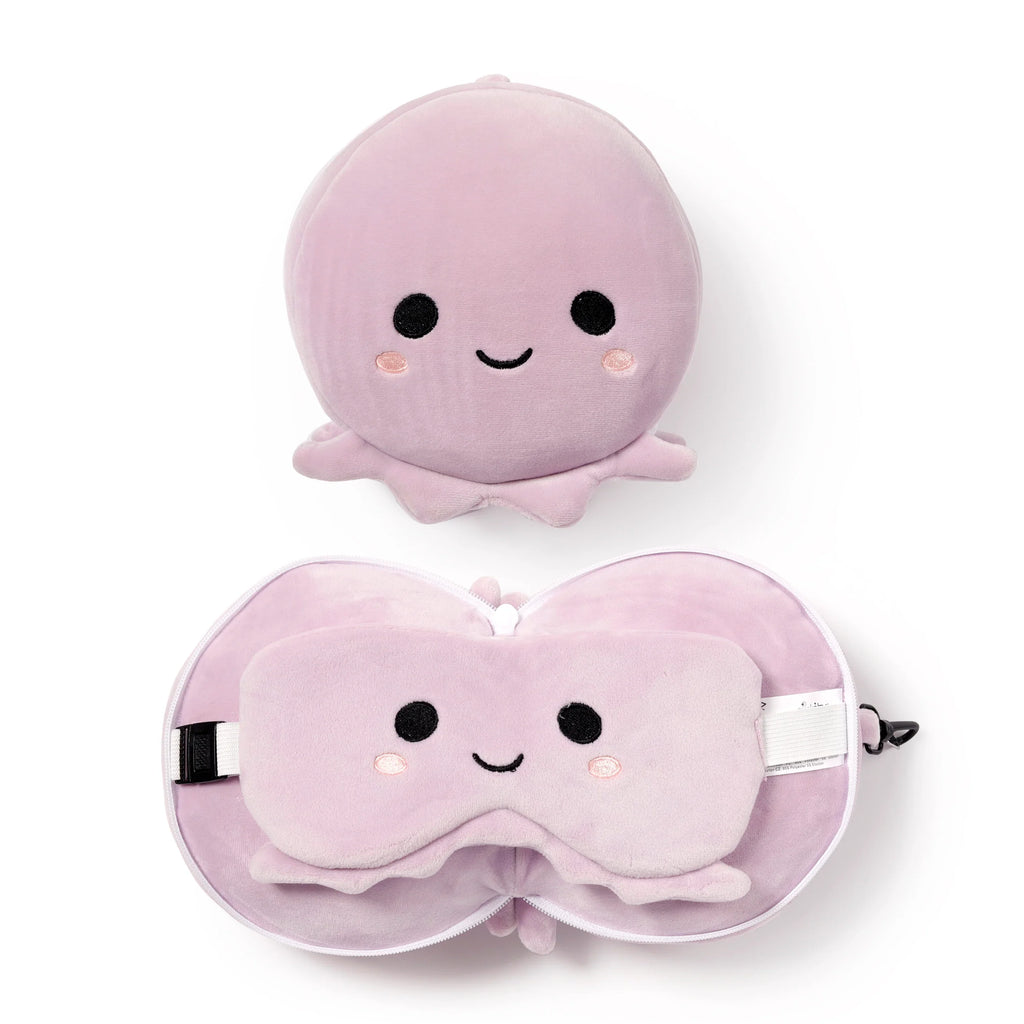 Flat lay overhead view of a lilac plush octopus toy and a matching travel pillow and eye mask set. The round plush octopus features embroidered black eyes, a smiling mouth, and pink blush cheeks. The U-shaped travel pillow is unzipped, revealing a coordinating eye mask with an identical embroidered face tucked inside. A white zipper and a black carabiner clip are visible on the pillow.