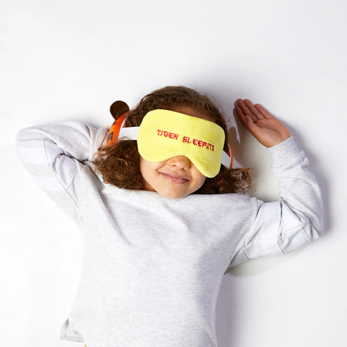 Lifestyle shot of a child wearing a yellow eye mask with red 'TIGER SLEEPING' text, alongside an orange and brown tiger plush travel pillow, resting on a white surface.