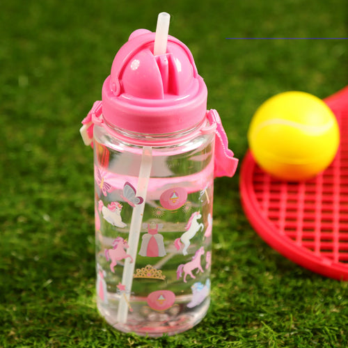 In-context lifestyle shot of a children's clear water bottle with a pink lid, pop-up straw, and pink carrying strap. The bottle displays pink and purple unicorn, butterfly, and princess designs, sitting on green artificial grass, with a blurry red tennis racket and yellow ball in the background.