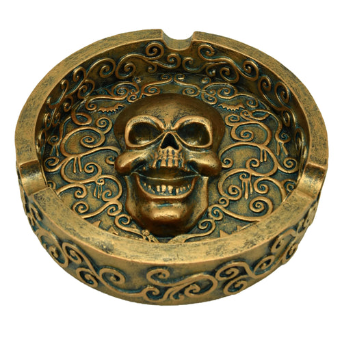 Overhead view of a decorative round ashtray with a metallic brushed gold effect finish, featuring a central grinning skull and intricate swirling patterns, with cigarette rests along the rim.