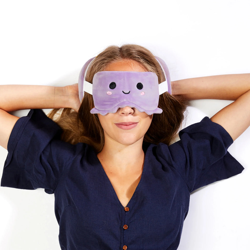 In-context lifestyle shot from above showing a woman lying down, wearing a purple plush octopus eye mask with a smiling embroidered face and pink cheek details, with parts of a matching purple travel pillow visible around her head.